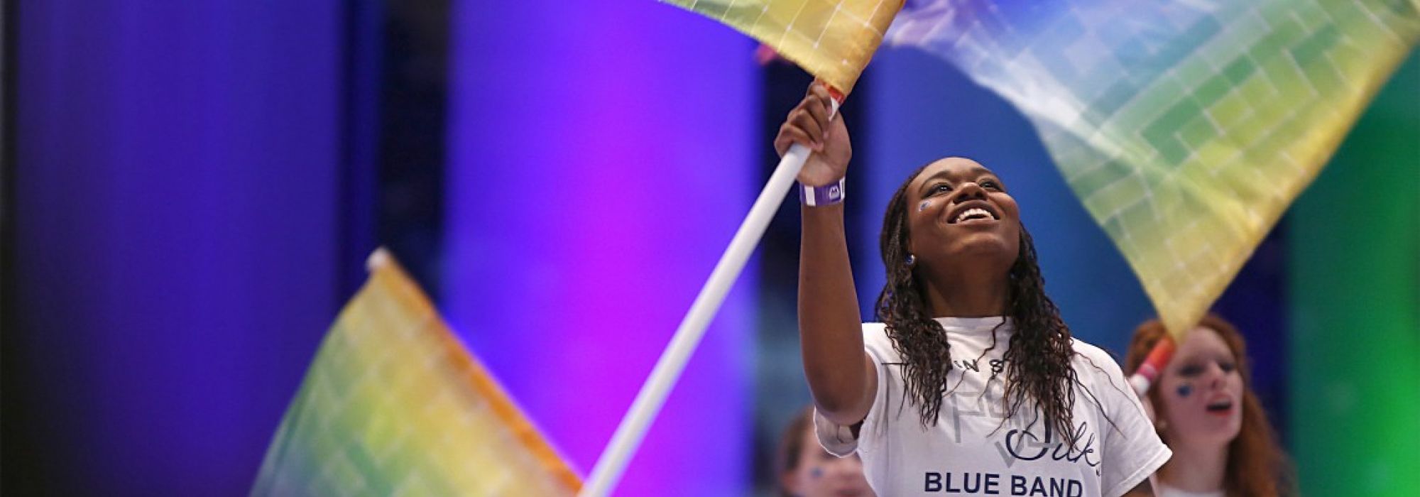 A young black student wearing a white t-shirt with a Blue Band Silks logo spins a rainbow-colored flag in the air.