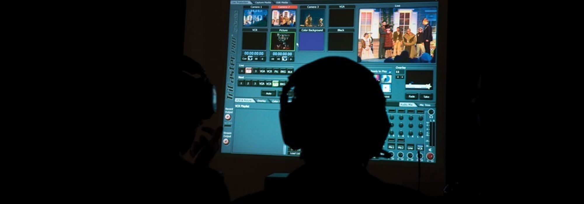 Two students in a control booth silhouetted in front of a monitor producing a live webcast of a theatre performance.