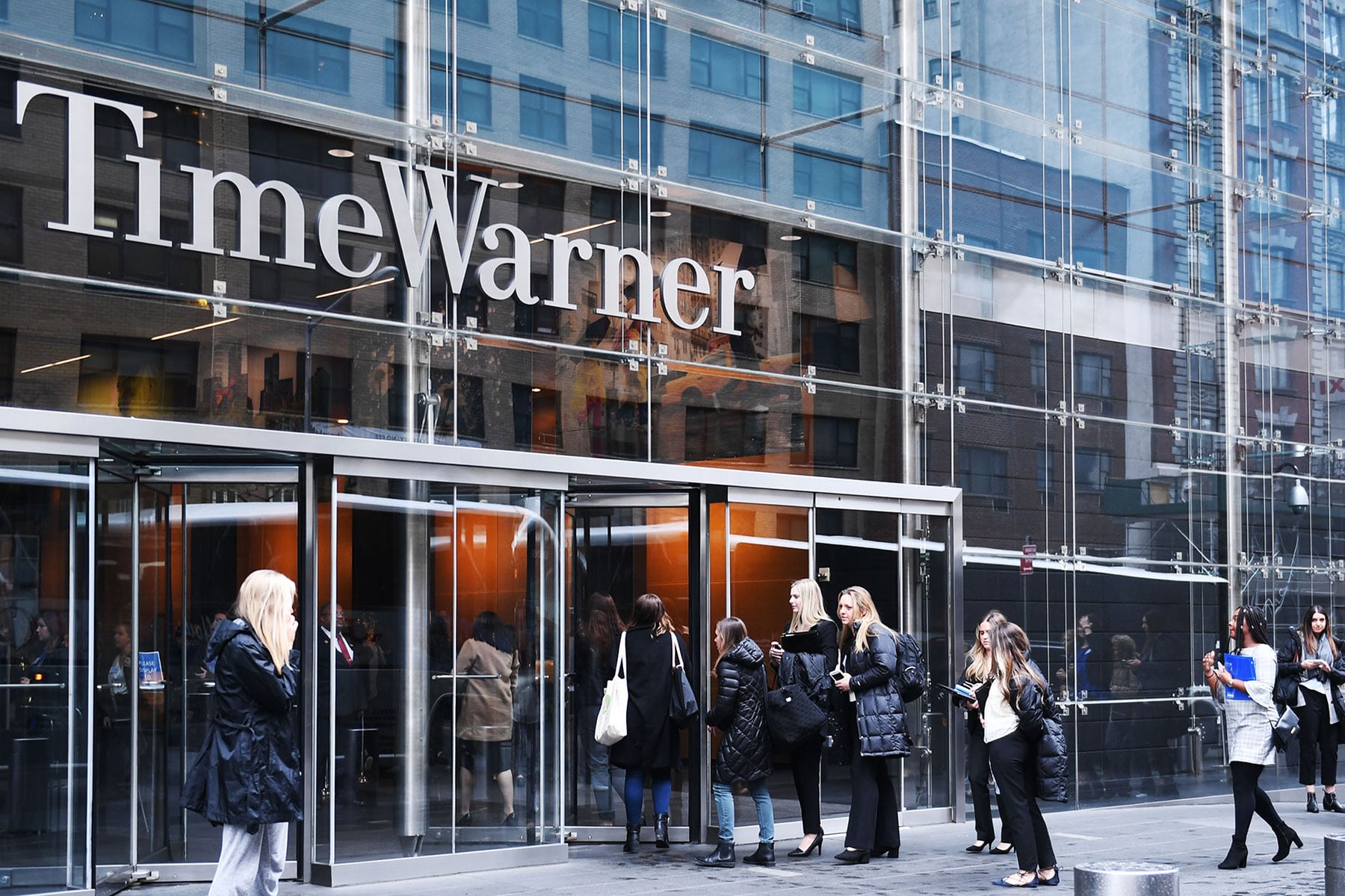 A number of young women dressed in winter clothes, file into an office building with the Time Warner logo above the door