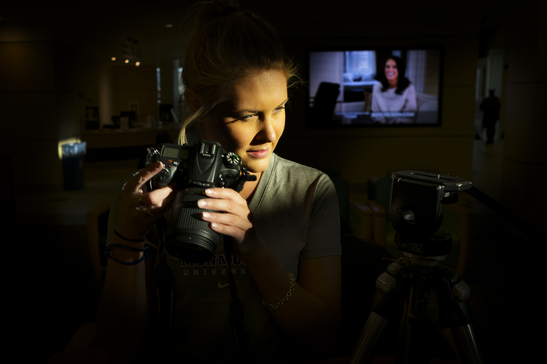 A young woman's face and camera are lit by stylish light with a very dark, moody background