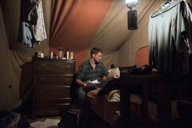 Zack sits at a desk in his tent, arranging school papers.