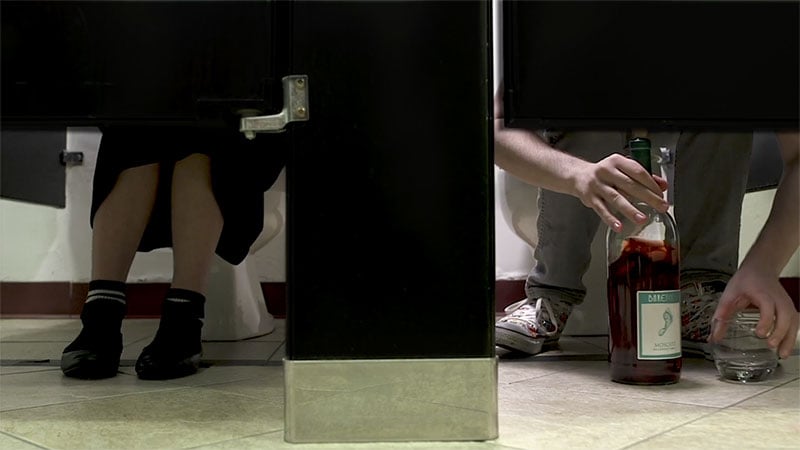Two pairs of legs are seen beneath the door of bathroom stalls. The person on the left has a bottle of wine near their feet.