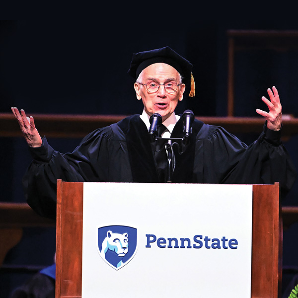 
	
Bellisario Alumnus Don Roy King speaks at a commencement ceremony with arms spread wide and hands up. He is wearing blue commencement robes and a six-sided hat and stands behind a podium with a Penn State logo on its front side.