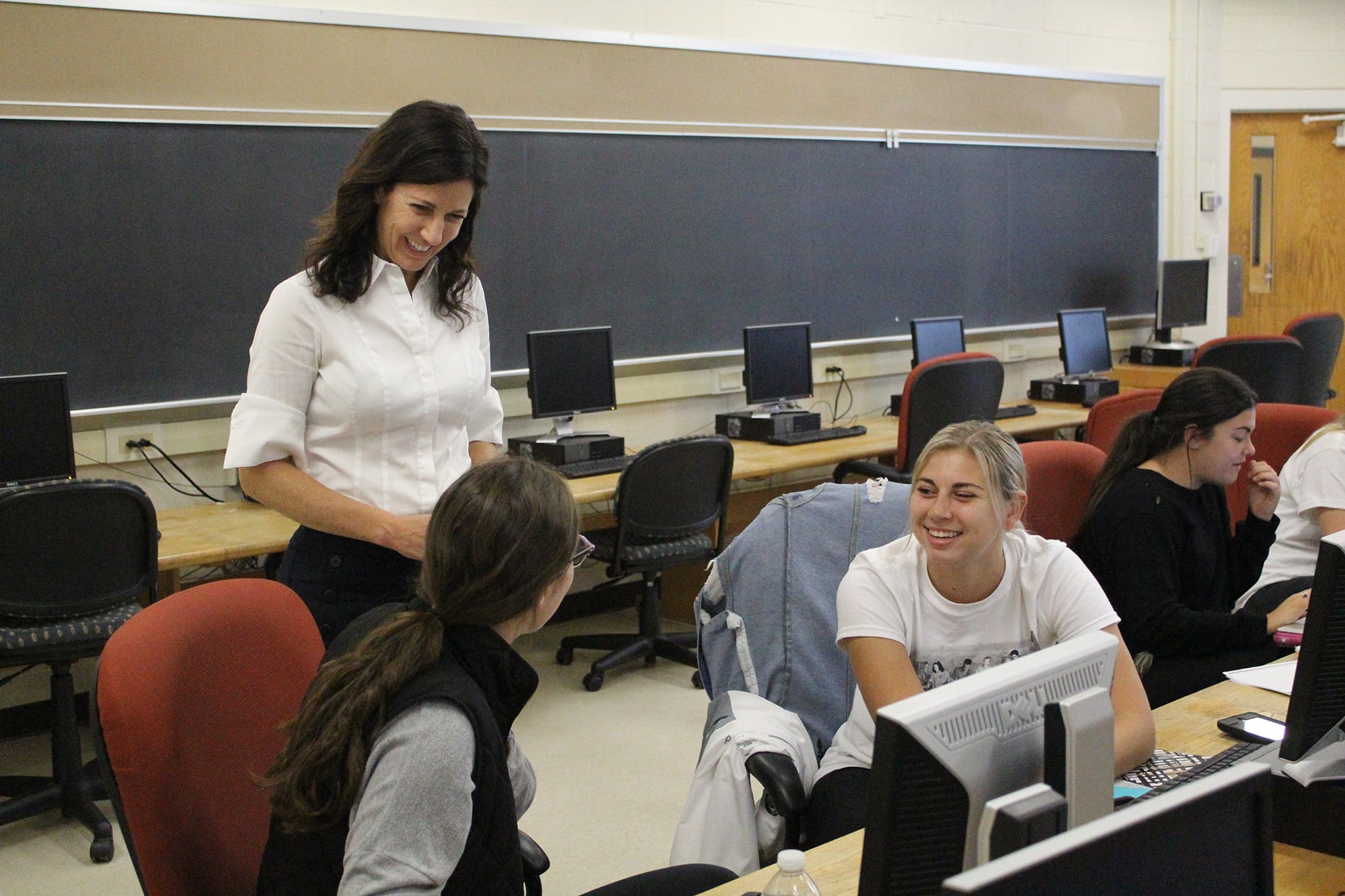 Tara Wyckoff smiles broadly while talking to two students sitting behind a computer monitor during a discussion. Wyckoff has long brown hair and is wearing a white dress shirt.
