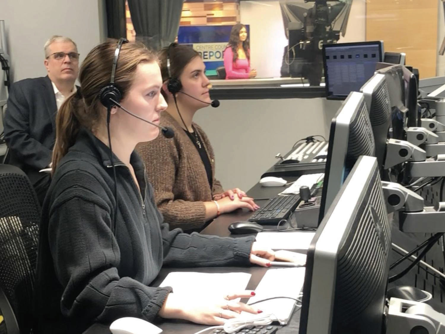 Students wearing headsets sit behind a television studio production desk working on a broadcast.