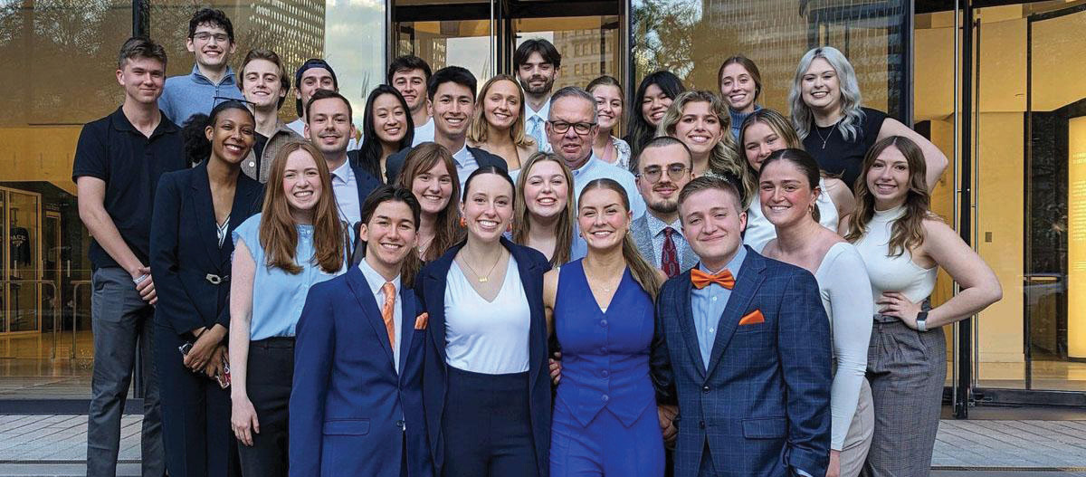 A group of students in smart, business casual dress pose in a building lobby.