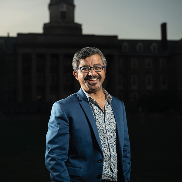 A man wearing a dark blue jacket and patterned shirt with graying goatee and hair poses in front of the dark silhouette of a building with a clock tower.