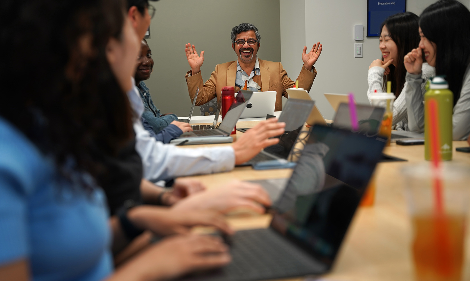 A faculty member with graying hair and goatee gestures broadly in a tan jacket and printed shirt at the head of a conference table with a group of graduate students.