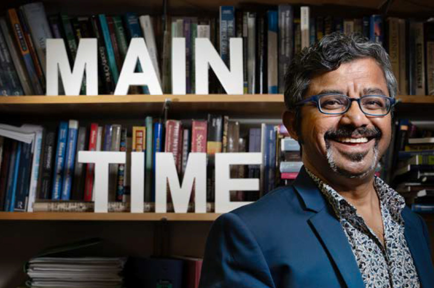 A man wearing a dark blue jacket and patterned shirt with graying goatee and hair poses in front of a bookshelf with cut out white letters that spell out Main Time