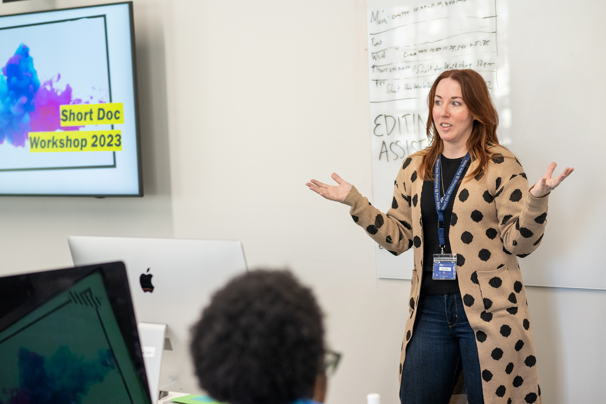 A woman in a long tan and black polka dot sweater gestures to a classroom with her arms out.