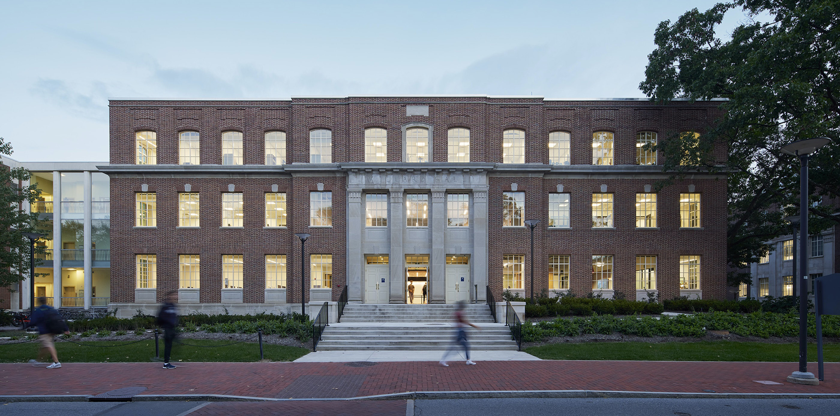 A subtle background photo of the front of Willard Building, a several story brick classroom building with four columns at the ntrance. Green lawn and bushes in the foreground.