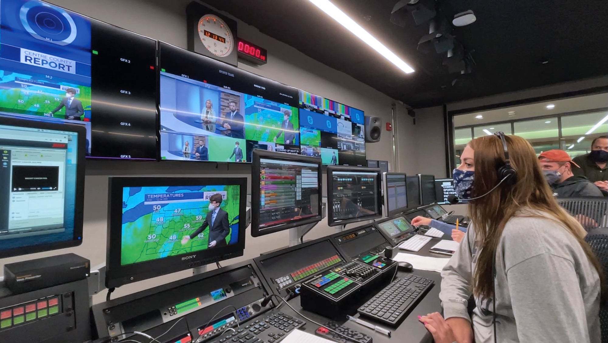 Students wearing masks during the COVID-19 pandemic sit behind a television studio production desk working on a live weather broadcast.