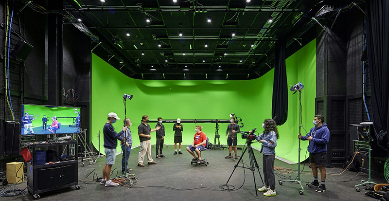 Wide screen look at severl students operating cameras in a big, open room with a green screen on the rear wall.