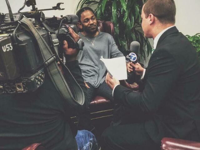 A student wearing a suit and holding a microphone interviews rapper Kendrick Lamar, sitting in a leather chair and wearing a gray shirt and slouchy beanie cap