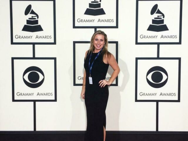 A student dressed in a black dress and wearing an ID badge, poses in front of a Grammy Awards backdrop