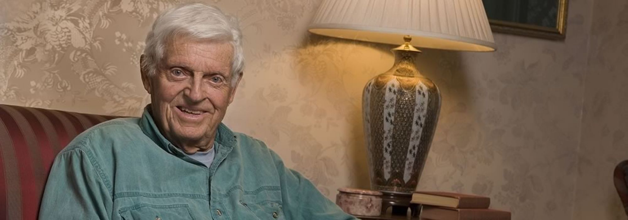 Alumnus Don Davis, smiling and wearing a green button down, sits on a couch with decorative wallpaper and lamp in the background.