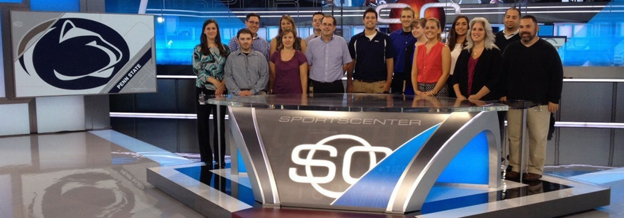 Fifteen members of the John Curley Center for Sports Journalism stand behind an anchor desk on the set of ESPN's SportsCenter