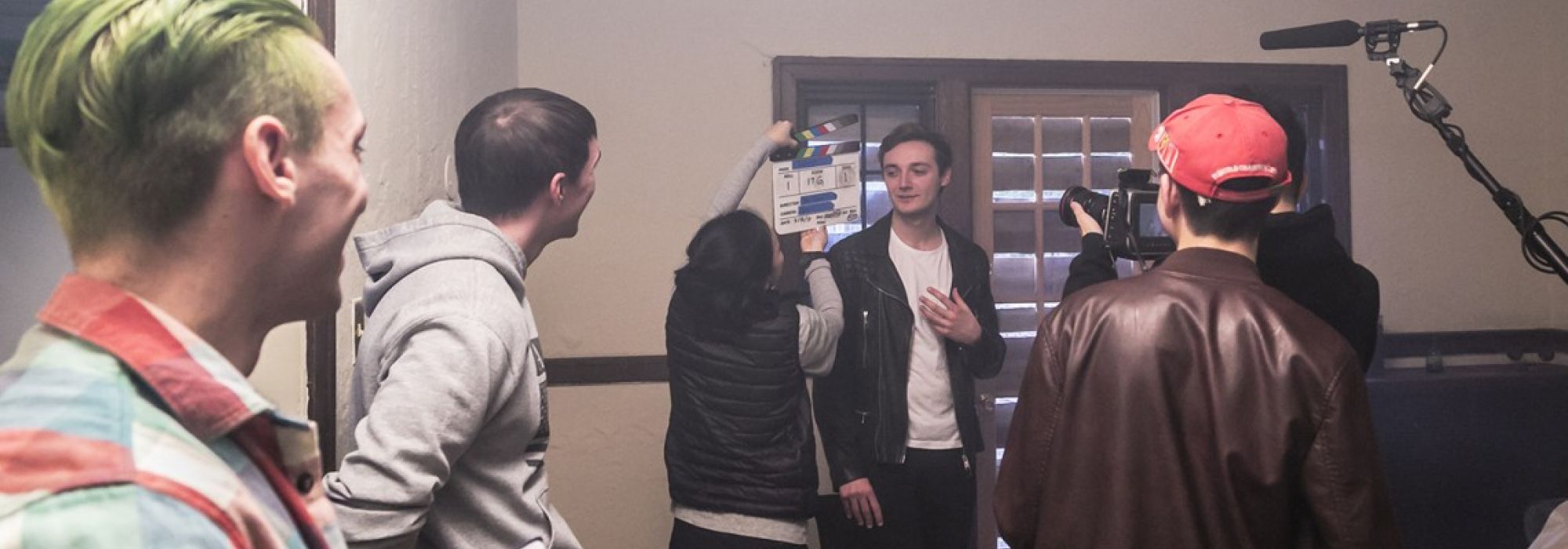 Six students on set of a film shoot. The focus is a student in white t-shirt and leather jacket who prepares to start acting as a producer stands in front of them with a clapperboard.