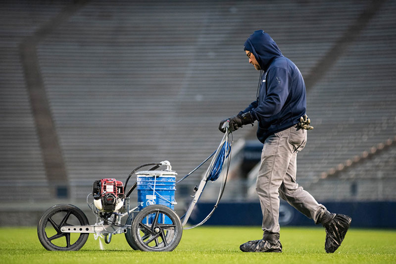 A groundskeeper in a blue hoodie and wearing protective coverings over his shoes uses a sprayer to paint yard lines on the Beaver Stadium grass.