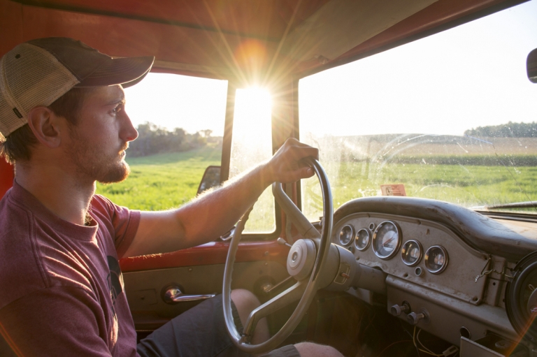 The sun shines brightly through the window as Zack drives his truck through a green farm field.