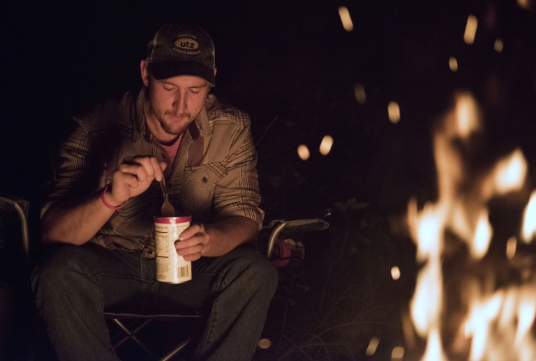 Zack sits in front of a campfire eating out of a can of beans with a spoon.