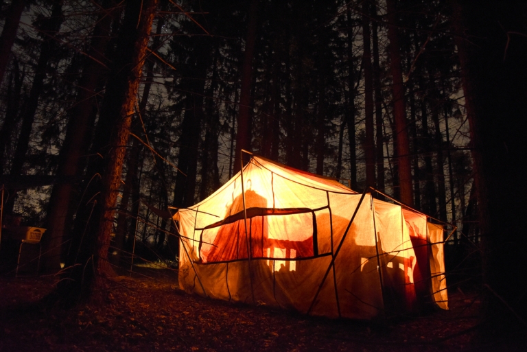 A canvas tent glows yellow from lighting inside with a stand of pine trees in the background.