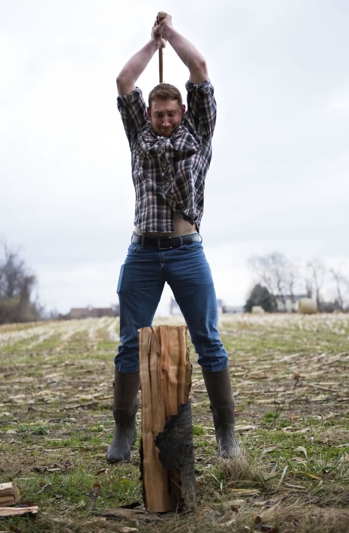 Zach Troyer, dressed in flannel, jeans, and boots swings an ax above his head to split a log.